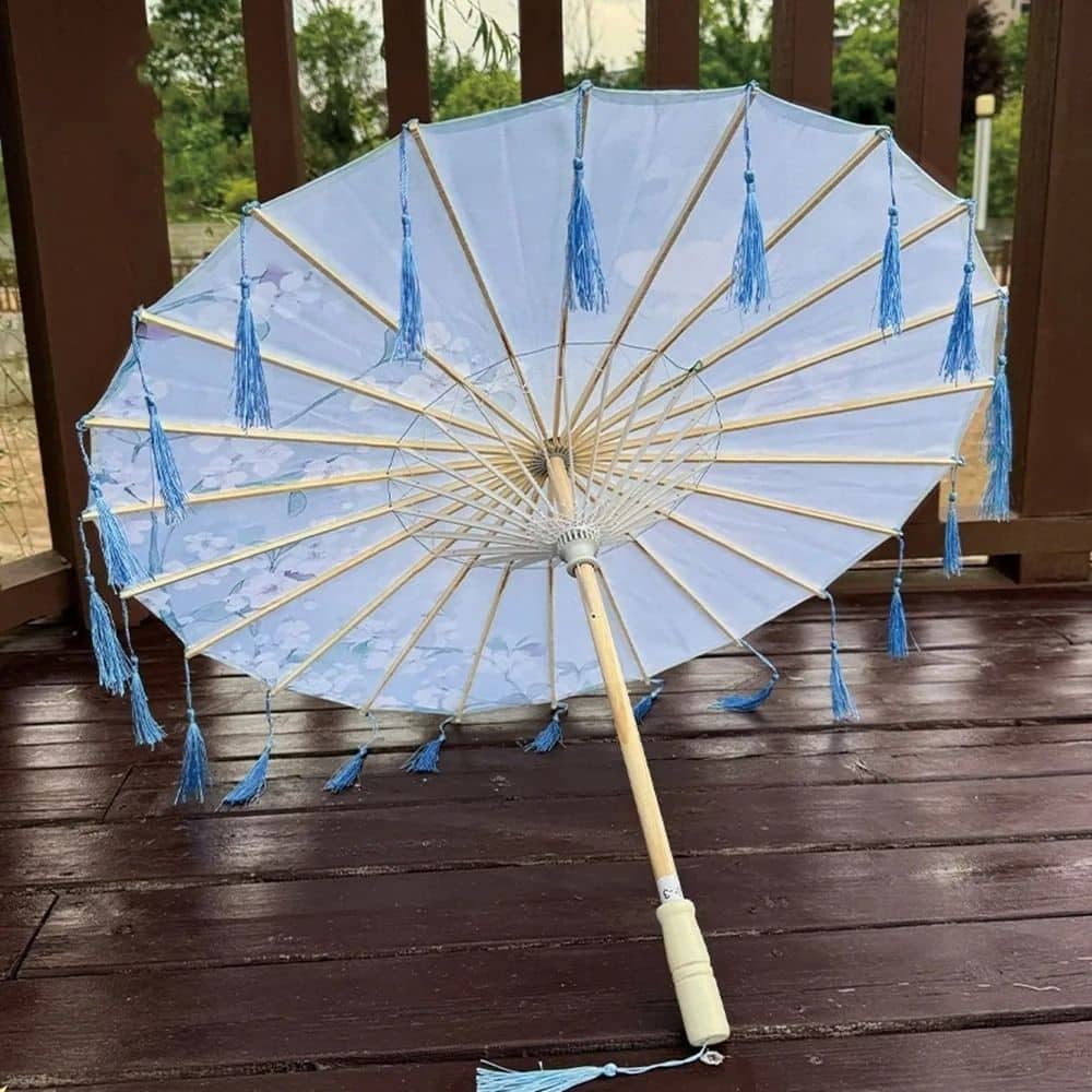 Ombrelle en papier bleu clair ornée de franges, posée sur un sol en bois, avec un décor naturel en arrière-plan