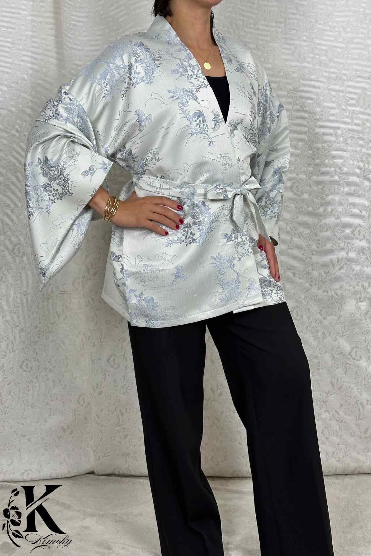 Femme en kimono argenté à motifs floraux bleus, posant avec une ceinture, sur fond texturé clair