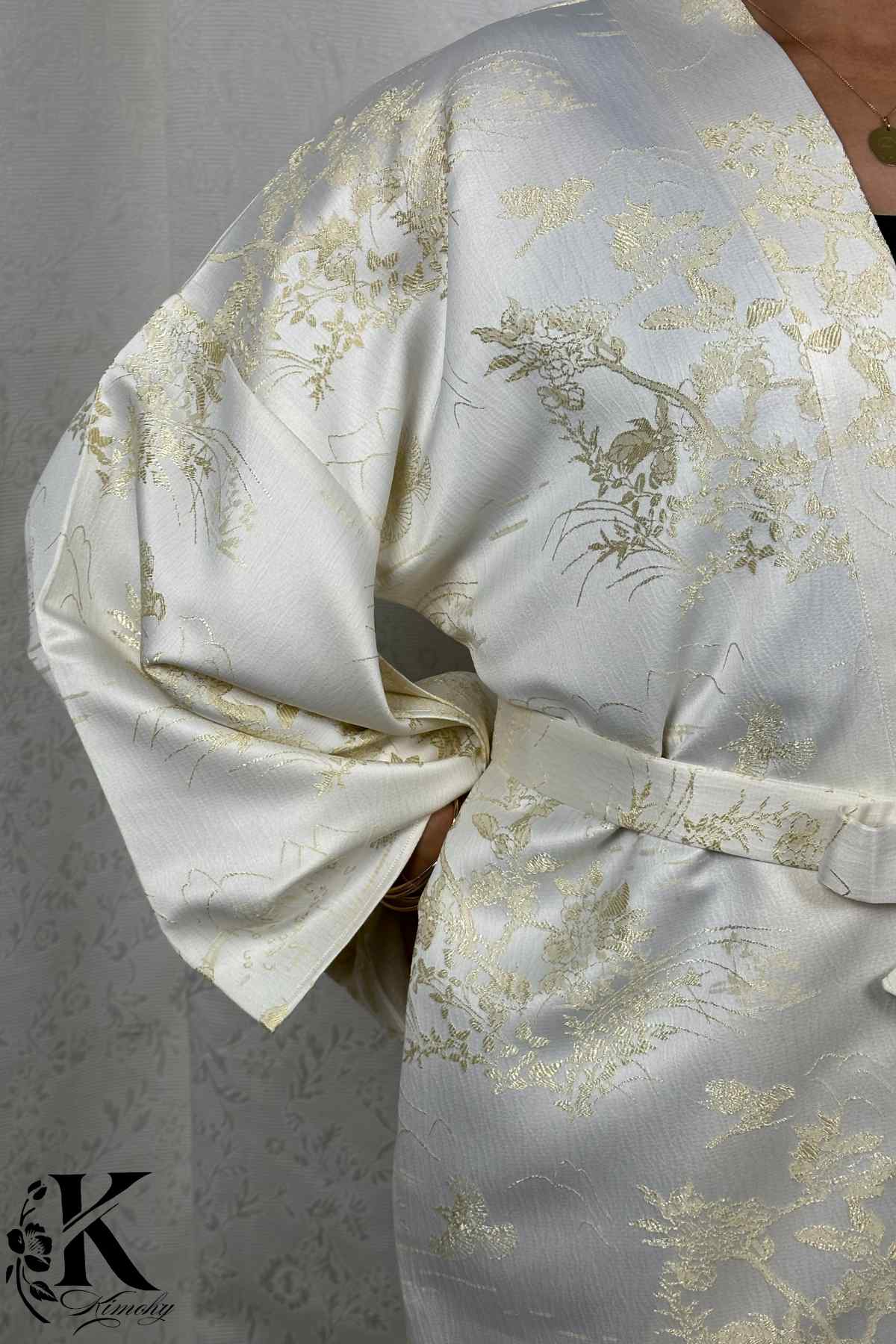 Kimono féminin en satin blanc avec motifs floraux dorés, ceinturé à la taille, posé sur un fond texturé