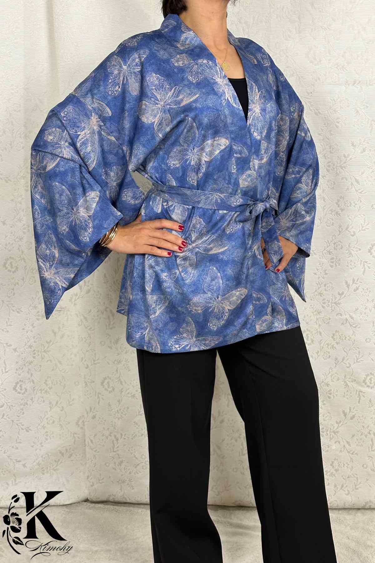 Femme portant un kimono bleu à motifs de papillons, avec des manches larges et un pantalon noir, sur fond clair