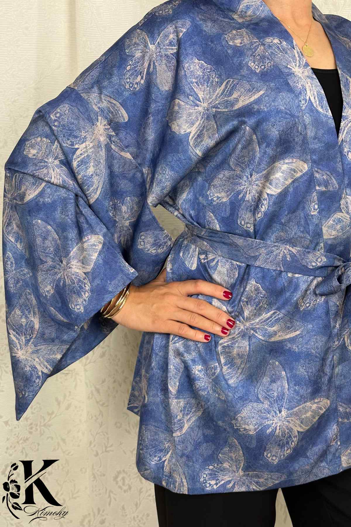 Kimono bleu avec motifs de papillons, manches larges, porté avec un pantalon noir, décor sobre en arrière-plan