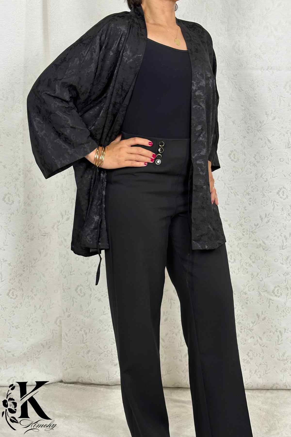 Femme en kimono noir avec motifs subtils, posant dans un décor clair et élégant, portant un pantalon noir