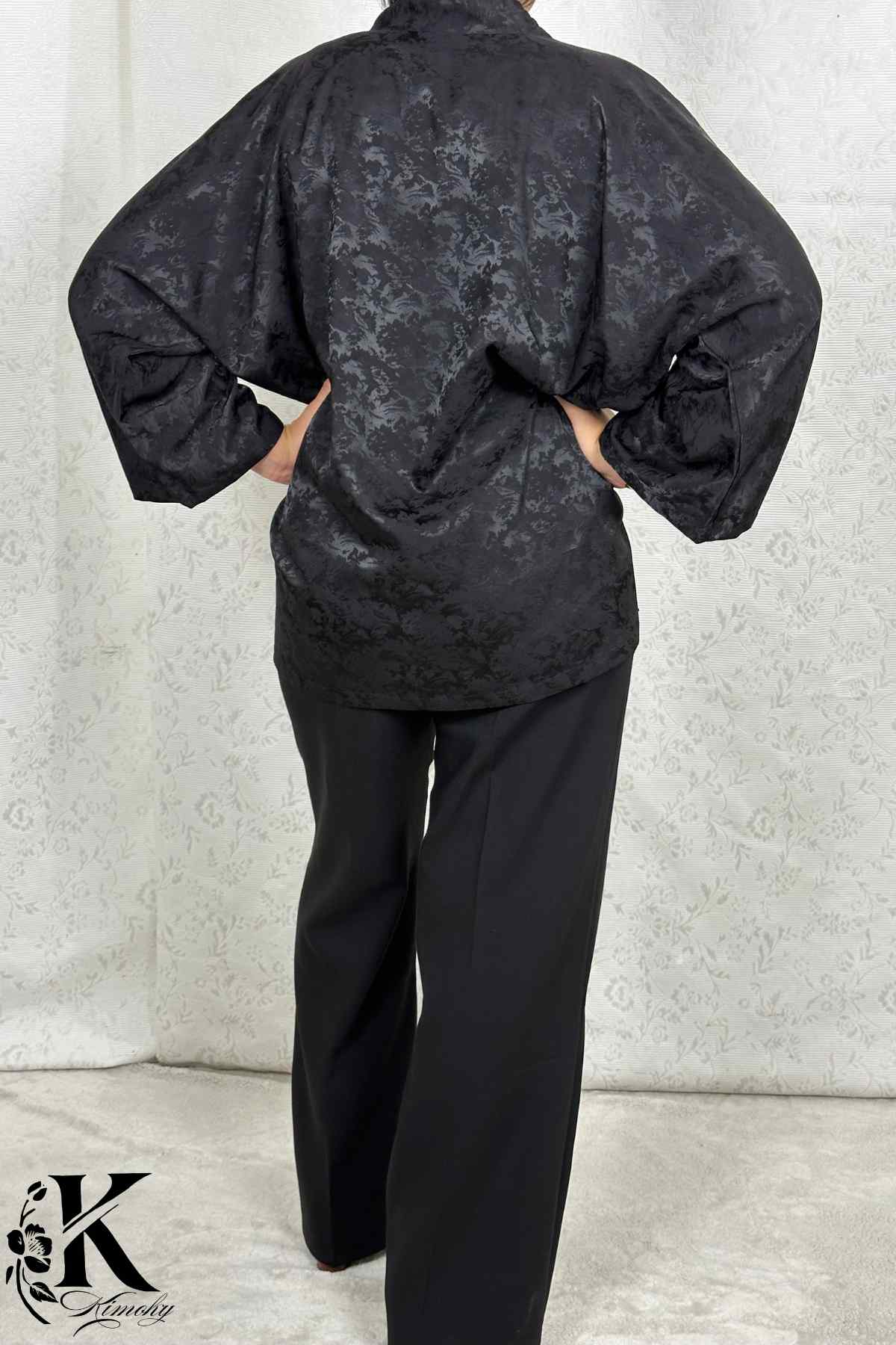Femme de dos portant un kimono noir à motifs, avec un fond de tissu clair et texturé
