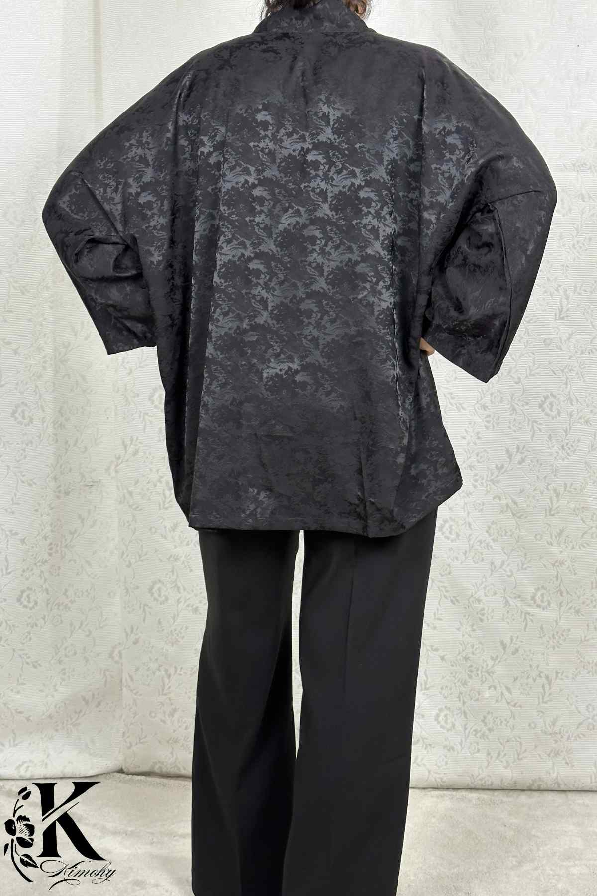 Kimono noir à motifs délicats porté avec un pantalon noir, sur fond clair et texturé