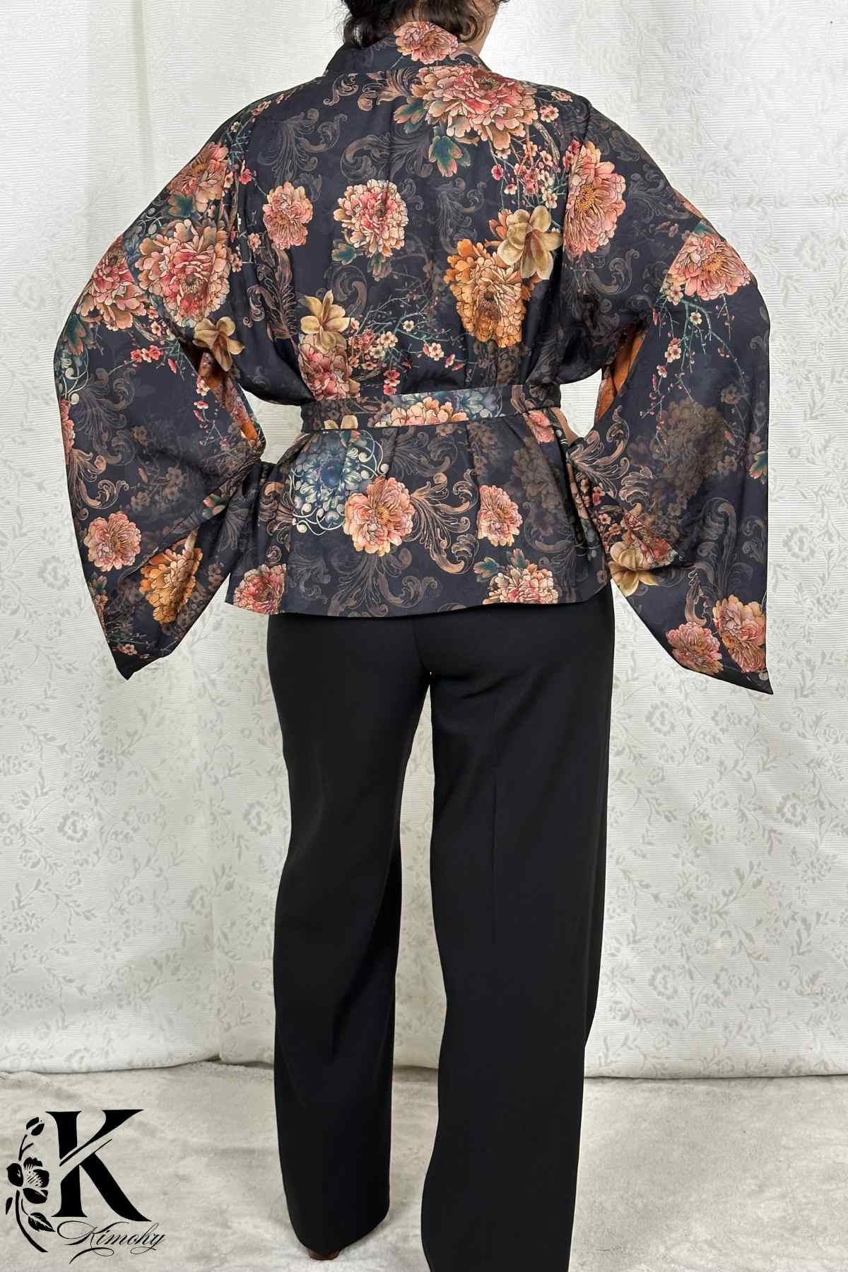 Kimono femme chic à fleurs roses sur fond noir, ceinturé, porté avec un pantalon noir dans un décor sobre