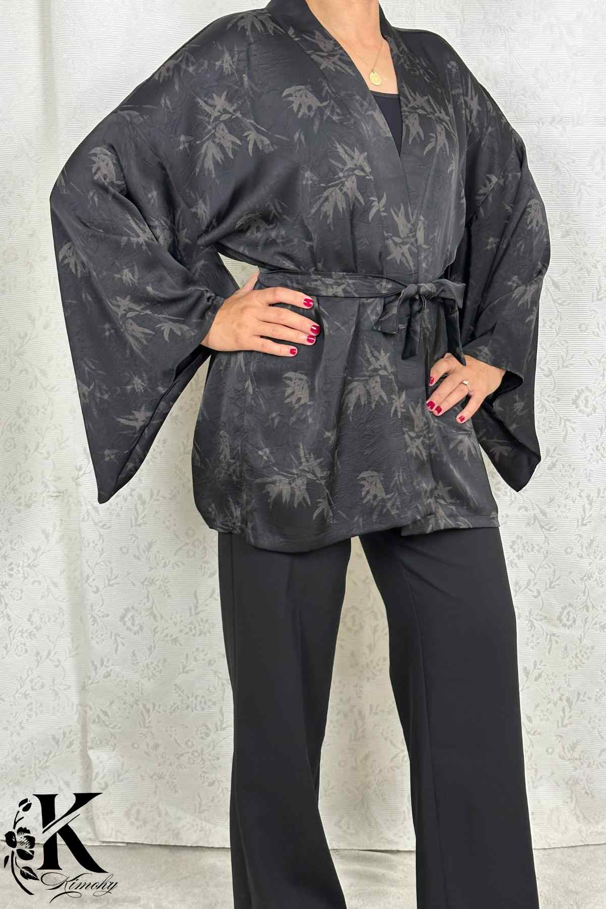 Femme en kimono noir à motifs de feuilles, posant dans un décor clair avec des pantalons noirs