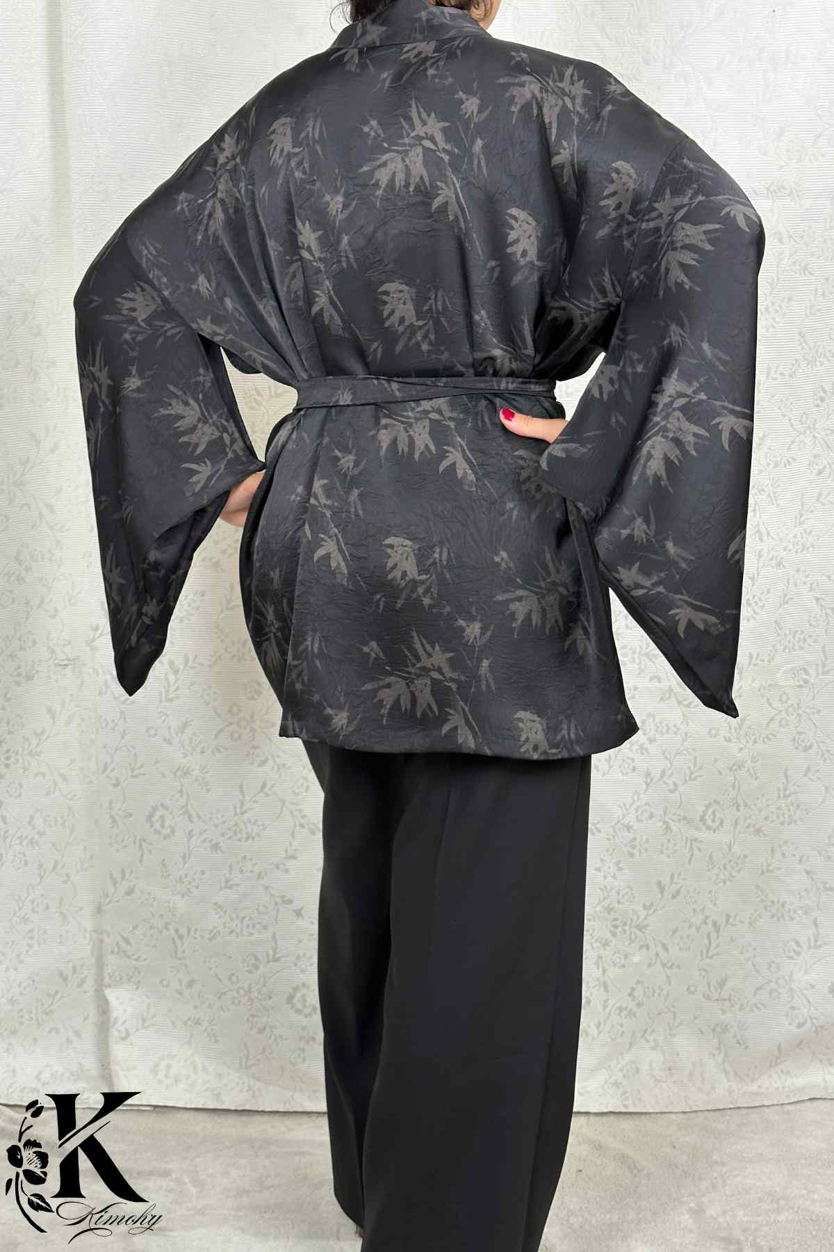 Femme portant un kimono noir avec motifs de bambous, dans un décor clair et texturé, avec des manches larges