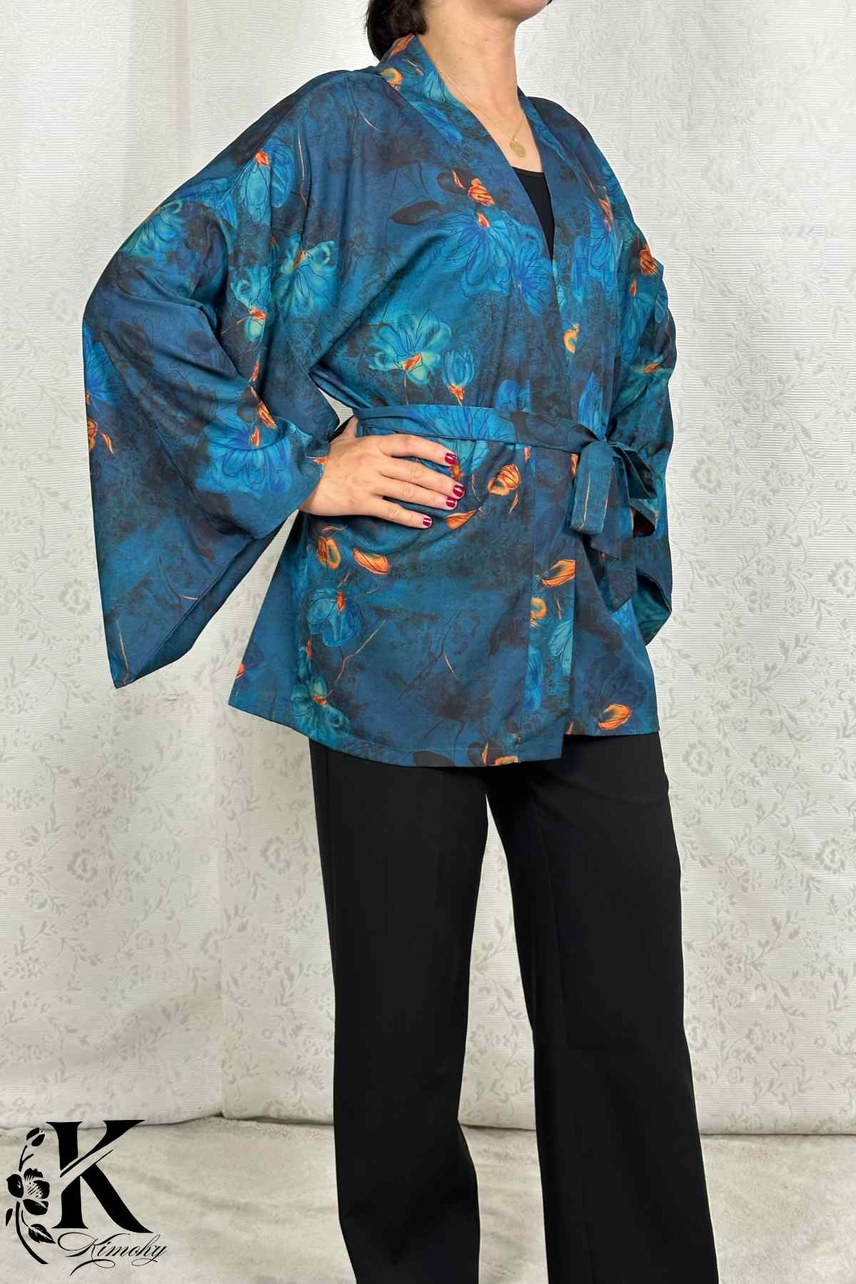 Femme en kimono bleu avec motifs floraux, posant dans un décor clair et texturé, ceinture nouée à la taille