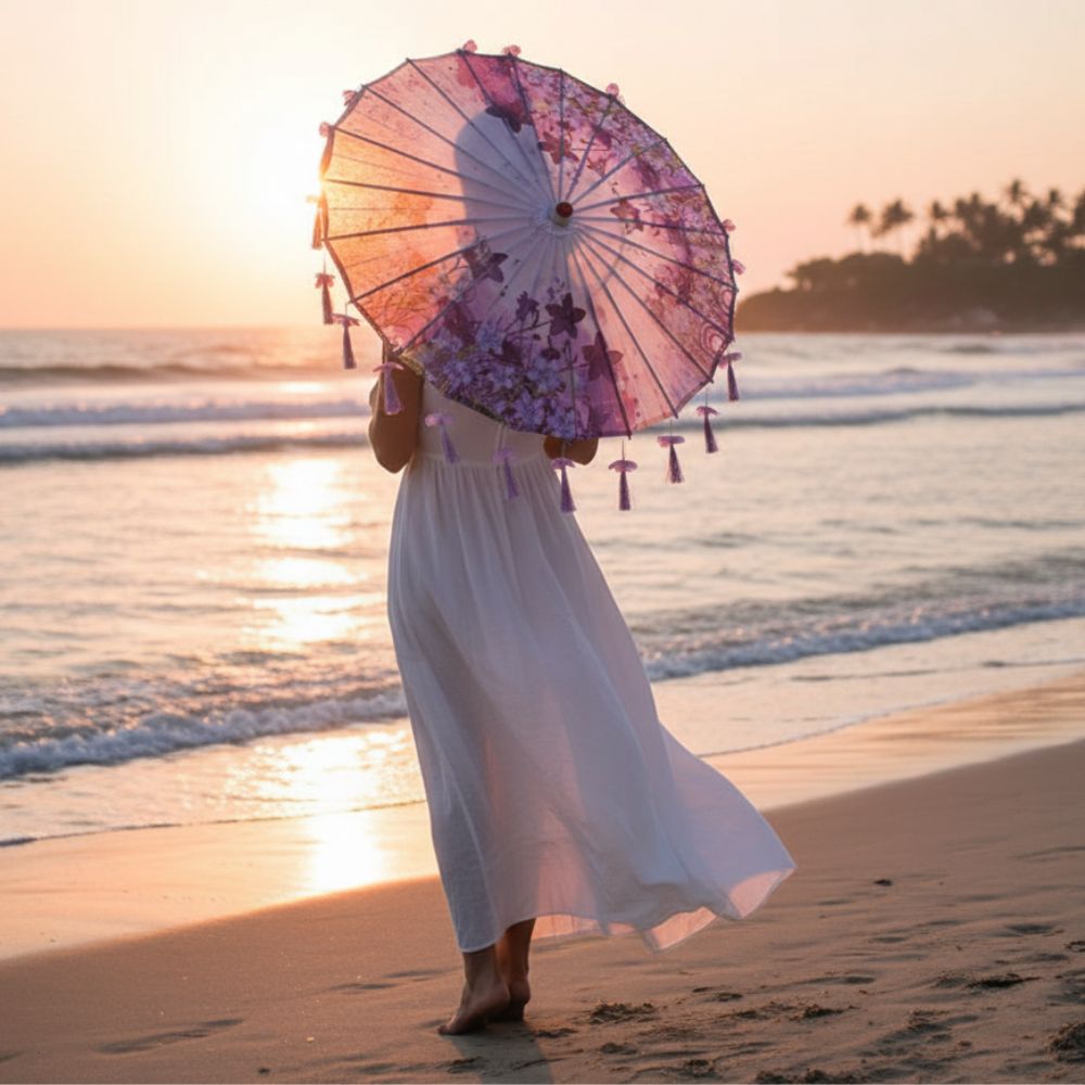 Femme en robe blanche tenant une ombrelle violette ornée de fleurs, marchant sur la plage au coucher du soleil