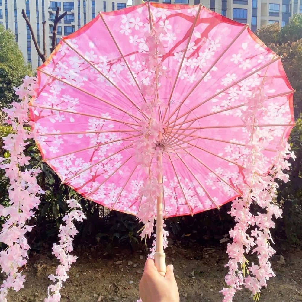 Parapluie japonais rose avec des motifs floraux, tenu à la main, entouré de verdure et d'un décor urbain en arrière-plan