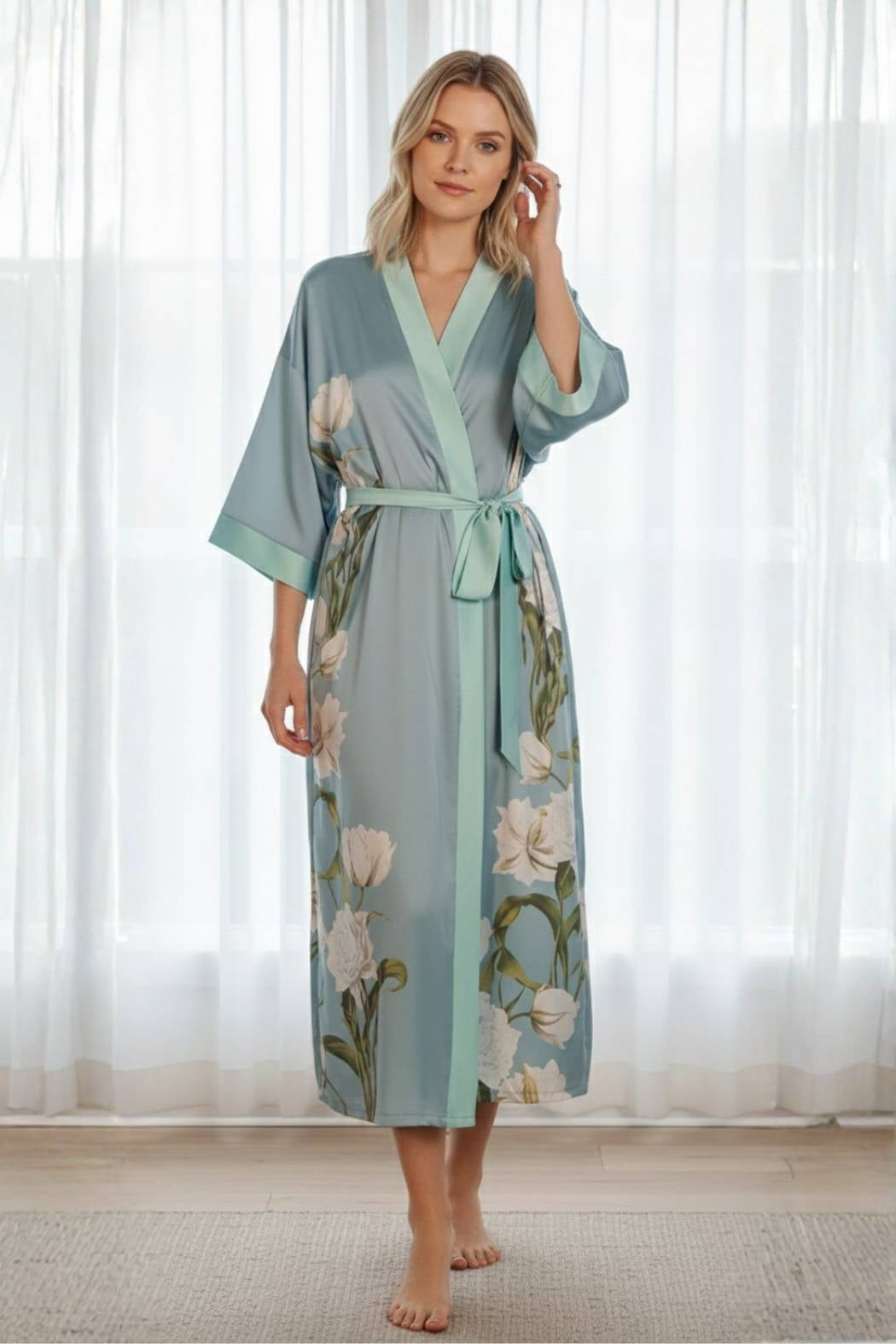 Kimono long en satin bleu clair avec des motifs floraux, porté par une femme debout, devant des rideaux blancs