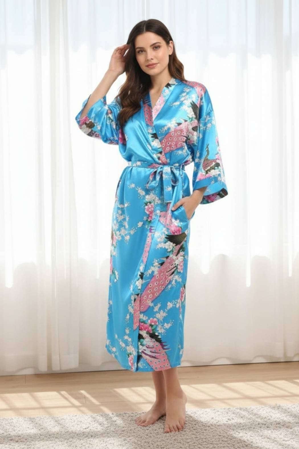 Femme en kimono bleu satiné orné de fleurs et oiseaux, posant dans une pièce lumineuse avec des rideaux blancs