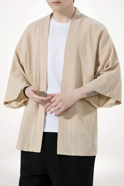 Homme portant un kimono beige en tissu léger, avec un t-shirt blanc et un pantalon noir, dans un décor minimaliste