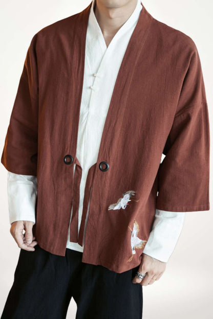 Homme portant un kimono brun avec des motifs de grues blanches, sur fond neutre, en posture décontractée