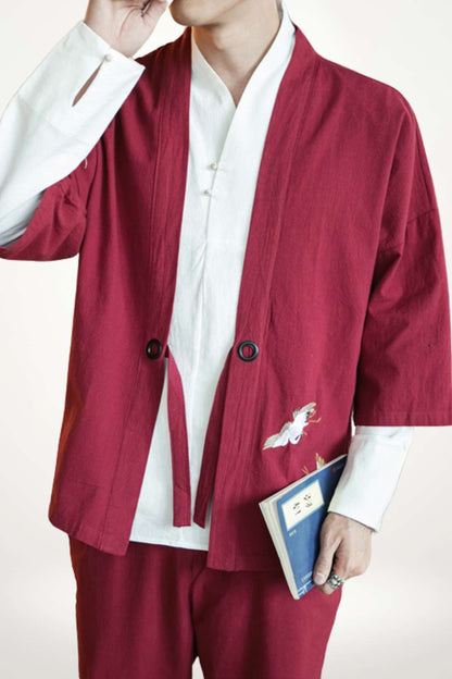 Homme portant un kimono moderne rouge avec des motifs brodés, tenant un livre dans un décor minimaliste