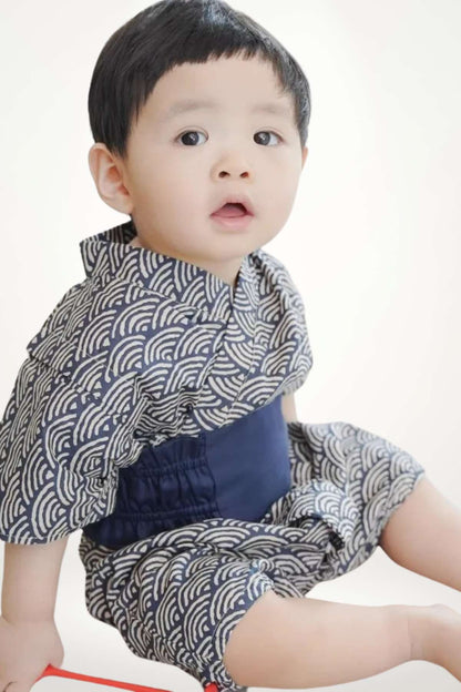 Un enfant assis porte un kimono bleu marine à motifs ondulés blancs, avec une ceinture bleue