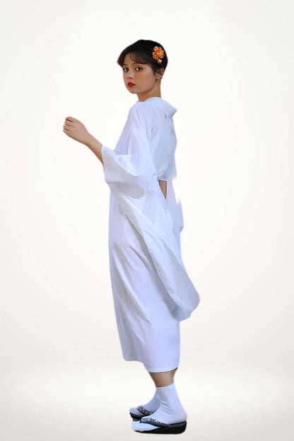 Femme en kimono blanc, aux manches larges, posant avec un décor neutre et une fleur orange dans les cheveux