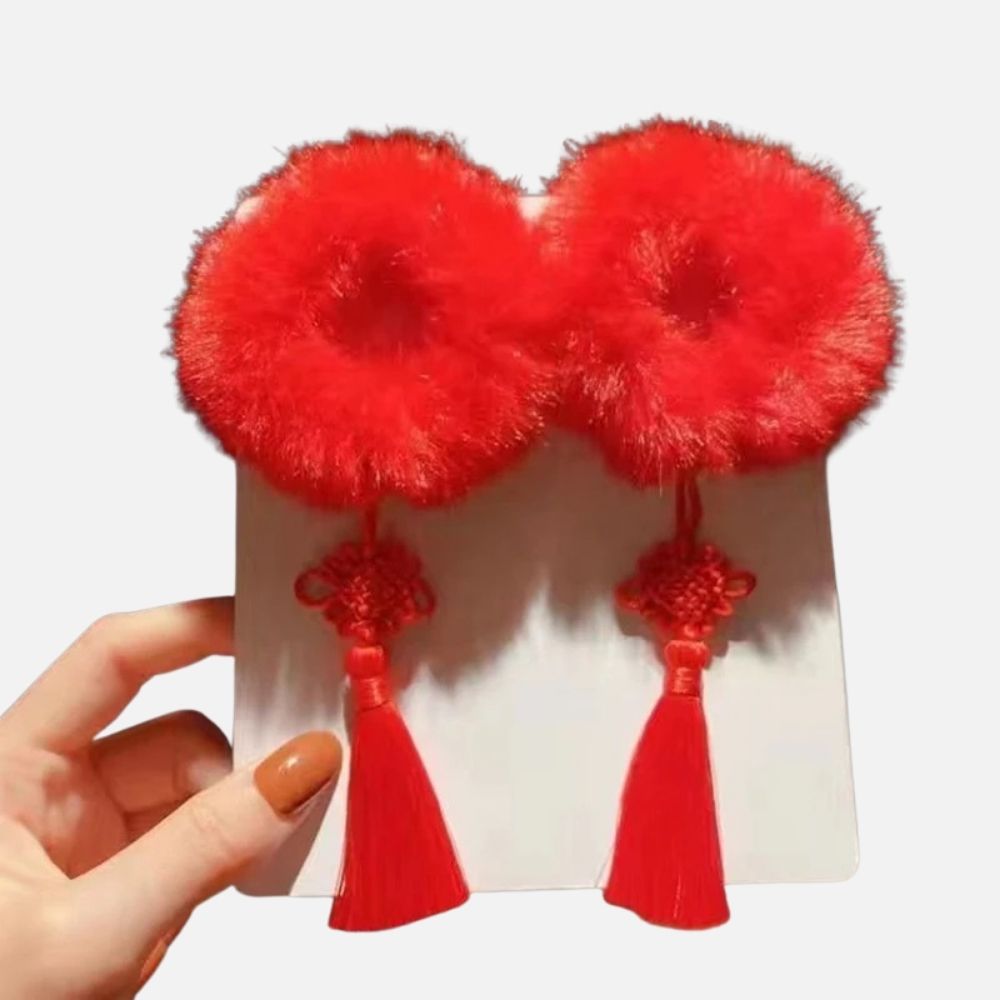 Deux accessoires rouges en fourrure avec des pompons et des franges, présentés sur un support blanc