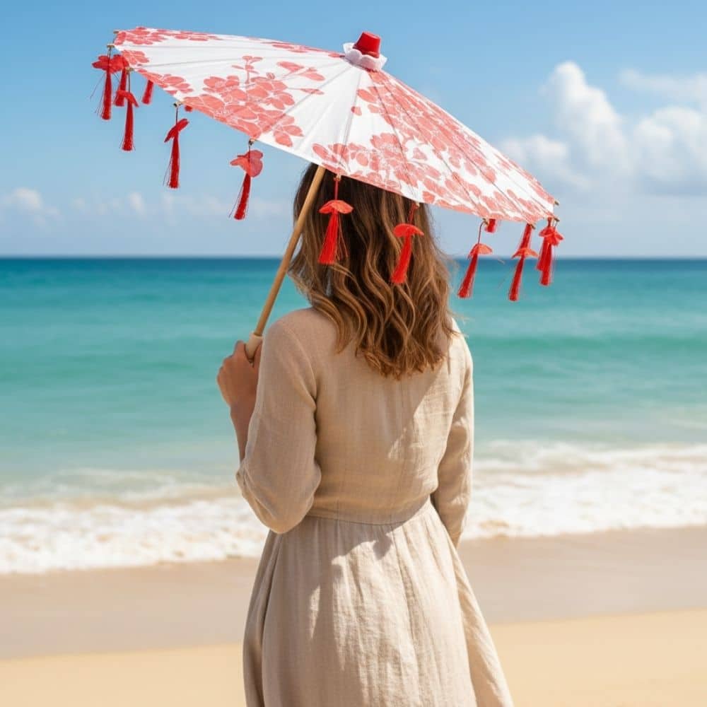 Femme de dos tenant une ombrelle rouge à motifs floraux sur une plage ensoleillée avec des vagues en arrière-plan