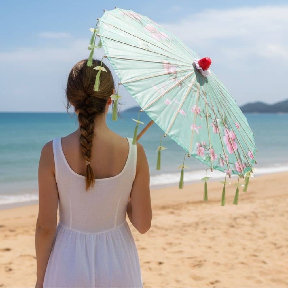 Femme de dos portant une robe blanche, tenant une ombrelle verte ornée de fleurs, sur une plage ensoleillée