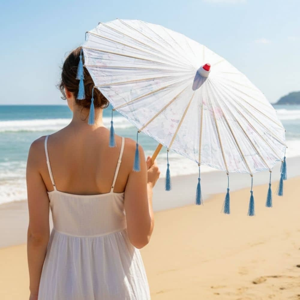 Une femme portant une robe blanche tient une ombrelle bleue à franges, face à la mer