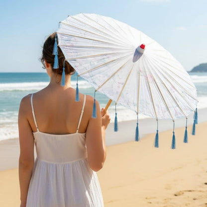 Une femme portant une robe blanche tient une ombrelle bleue à franges, face à la mer