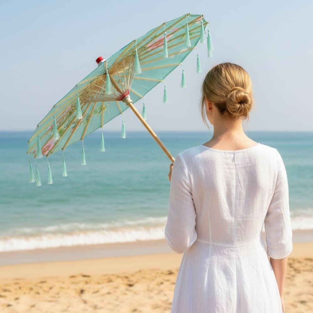 Femme en robe blanche tenant un parapluie bleu décoré de franges, face à la mer et au sable