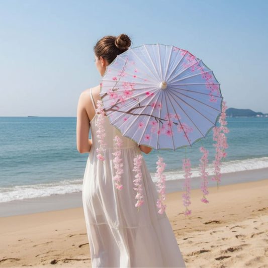 Femme de dos portant un parapluie japonais rose avec des motifs floraux, face à la mer et au sable