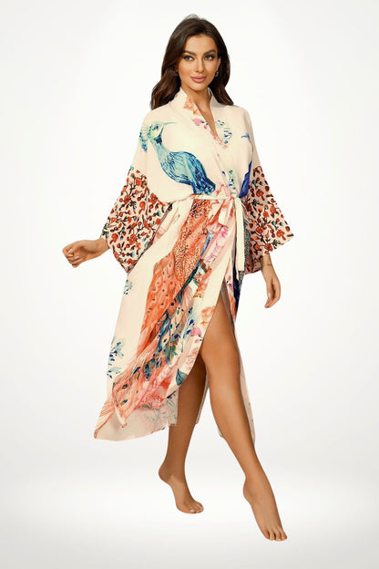 Femme en kimono satin aux motifs colorés de paons et fleurs, posant avec un sourire dans un décor minimaliste