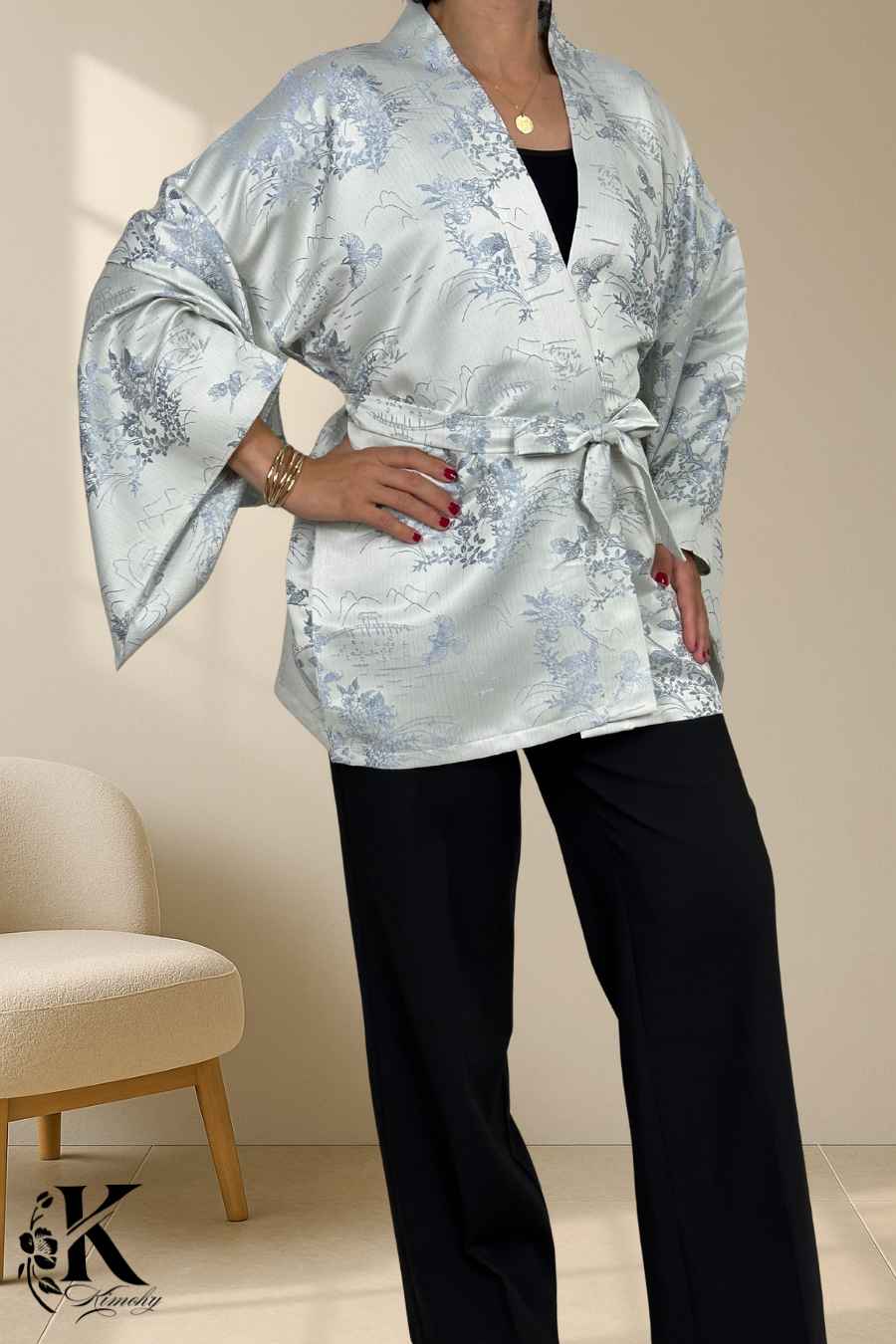 Femme en kimono argenté à motifs floraux bleus, posant avec une ceinture, sur fond texturé clair