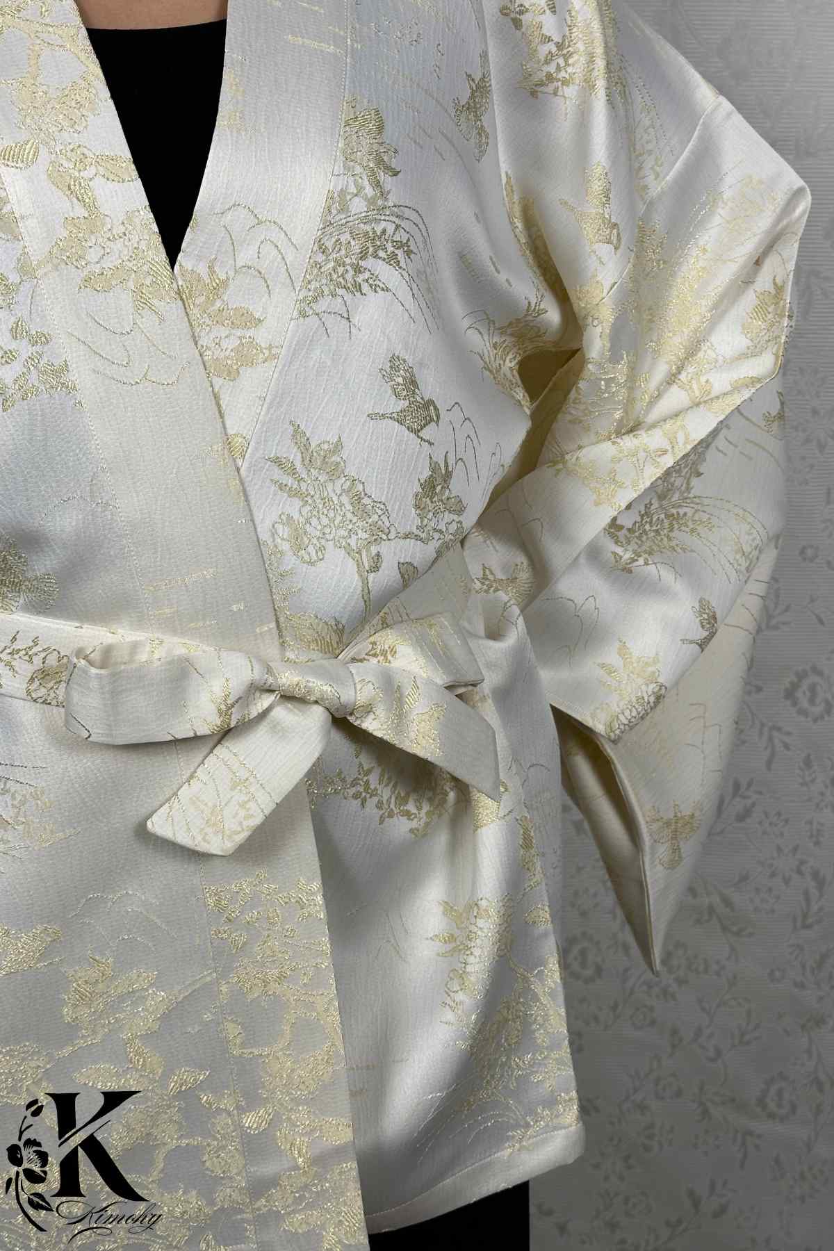 Kimono chic en satin blanc avec motifs dorés, noué à la taille, sur fond texturé clair