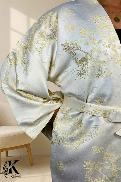 Kimono féminin en satin blanc avec motifs floraux dorés, ceinturé à la taille, posé sur un fond texturé