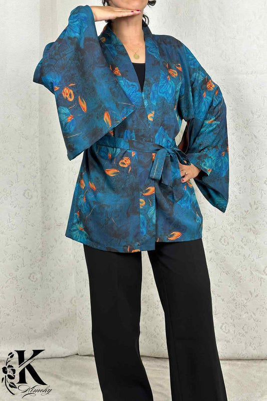 Femme en kimono bleu à motifs floraux orange, posant dans un décor clair et texturé, avec un pantalon noir