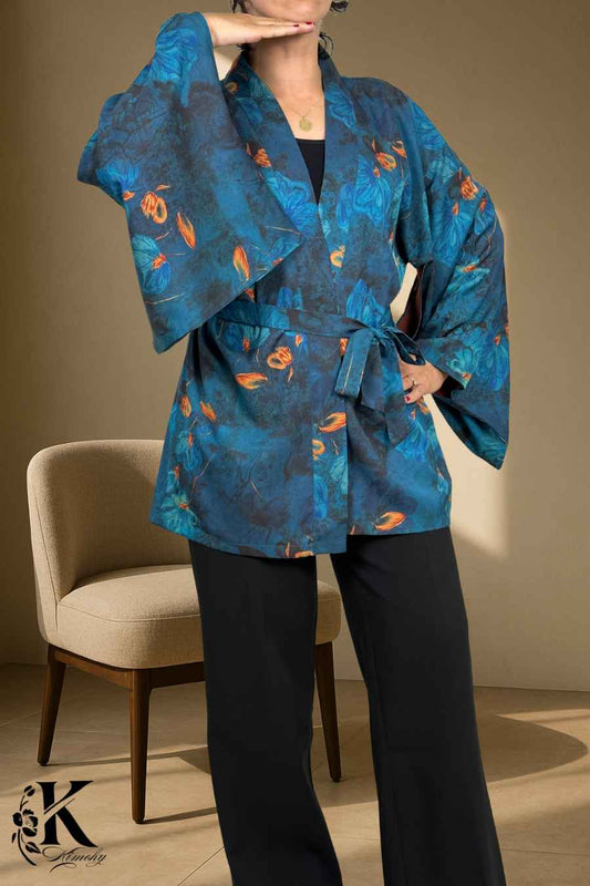 Femme en kimono bleu à motifs floraux orange, posant dans un décor clair et texturé, avec un pantalon noir