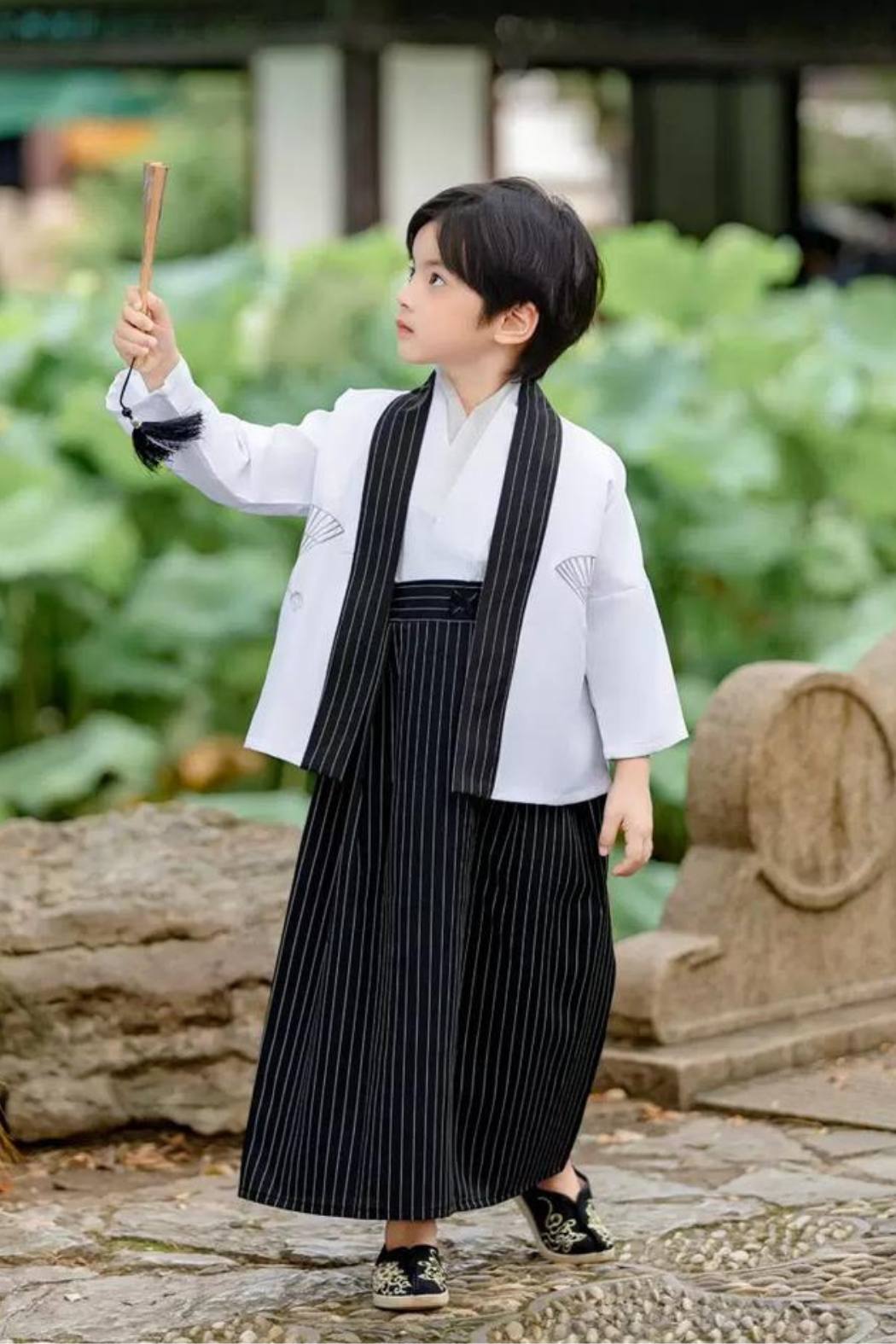 Enfant portant un kimono blanc avec des motifs, pantalon noir à rayures et chaussures noires, tenant un objet 