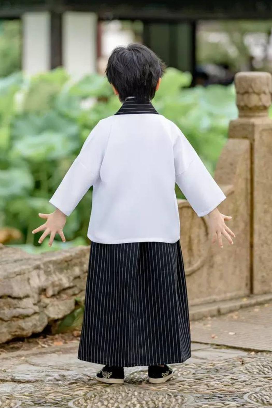 Enfant de dos portant un kimono blanc avec des motifs, pantalon noir à rayures et chaussures noires dans un jardin