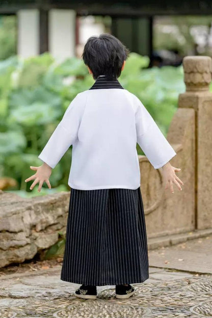 Enfant de dos portant un kimono blanc avec des motifs, pantalon noir à rayures et chaussures noires dans un jardin