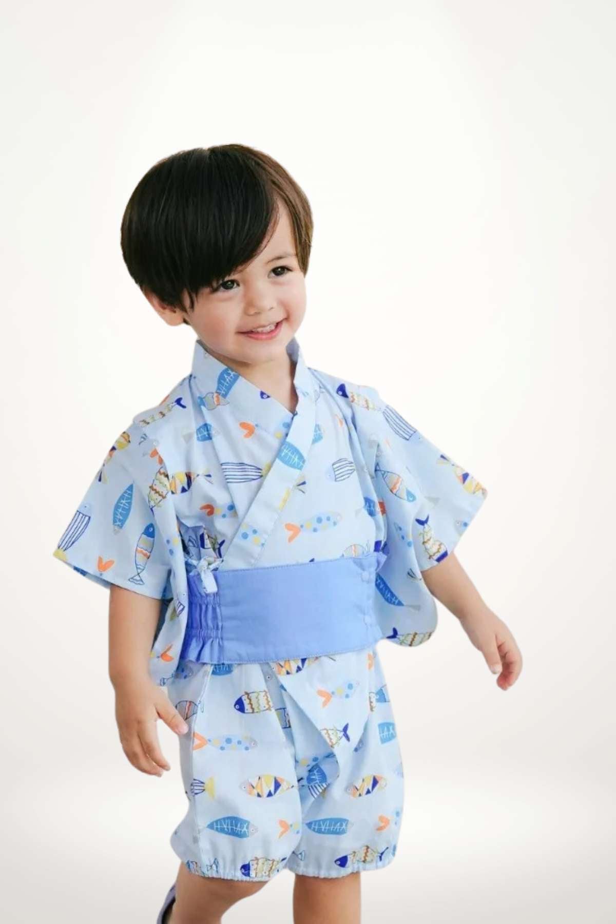 Un enfant souriant porte un kimono bleu avec des motifs de petits poissons, se tenant debout sur un fond clair