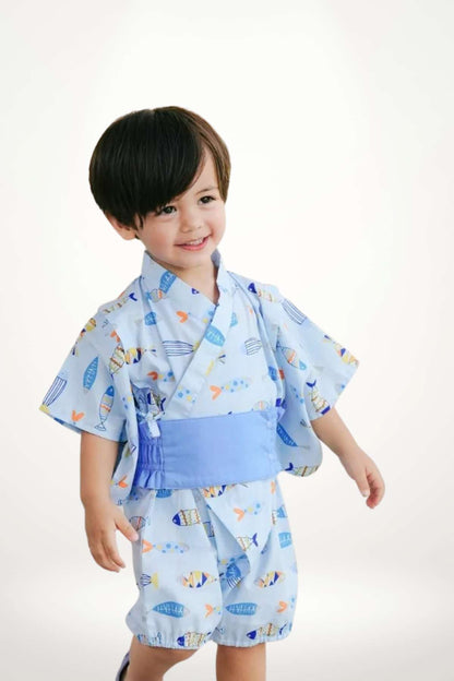 Un enfant souriant porte un kimono bleu avec des motifs de petits poissons, se tenant debout sur un fond clair