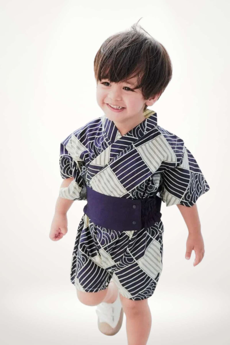 Un enfant souriant porte un kimono bleu et blanc à motifs géométriques avec une ceinture noire, sur fond clair