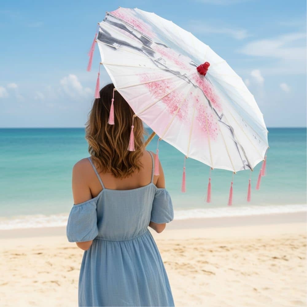 Femme en robe bleue tenant une ombrelle rose à franges, face à la mer sous un ciel bleu