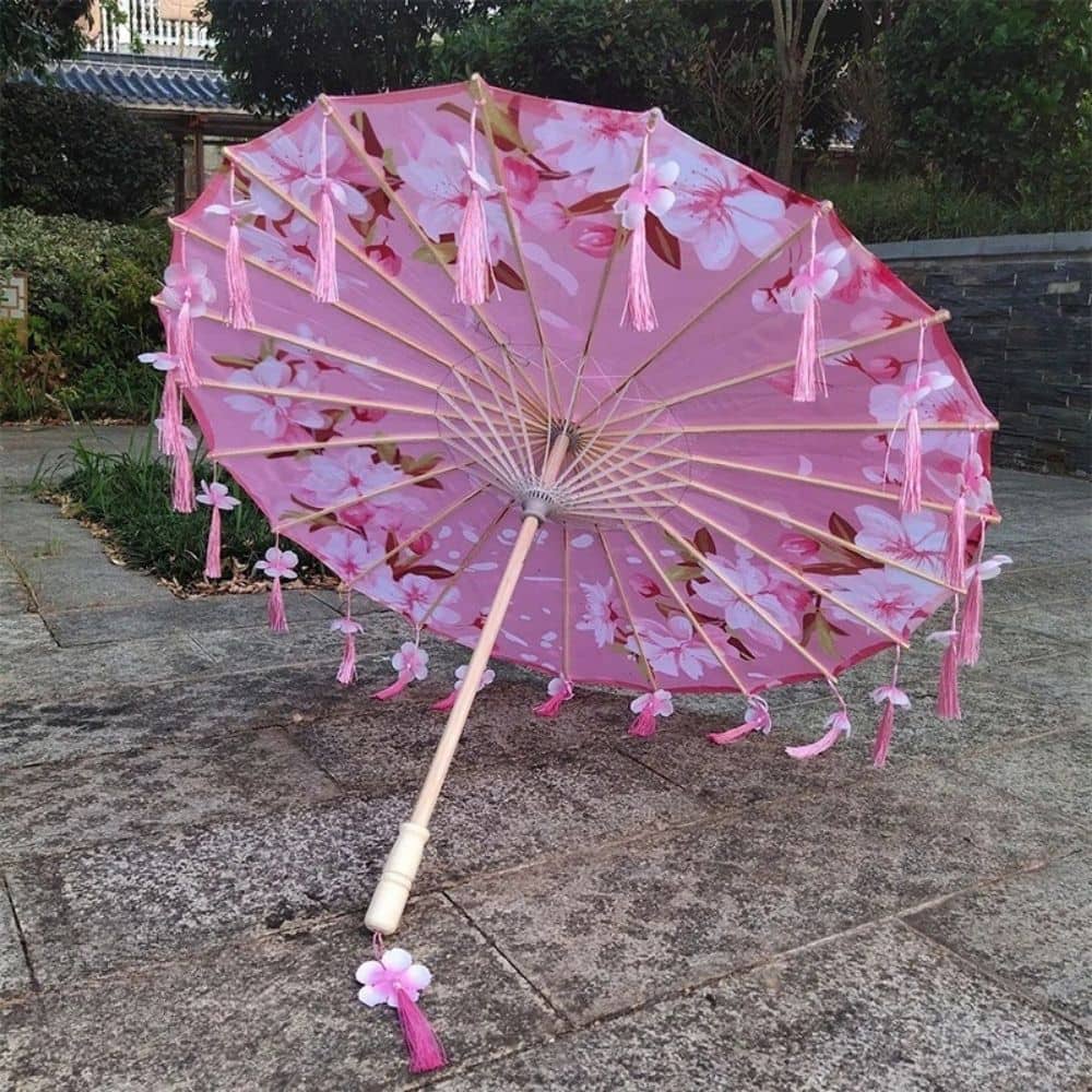 Ombrelle anti-UV 50 rose ornée de fleurs blanches et de pompons roses, posée sur un sol en pierre