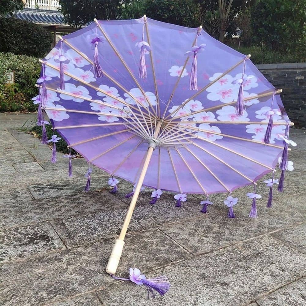 Ombrelle anti-UV adulte violette ornée de motifs nuageux et de franges, posée sur un sol en pierre