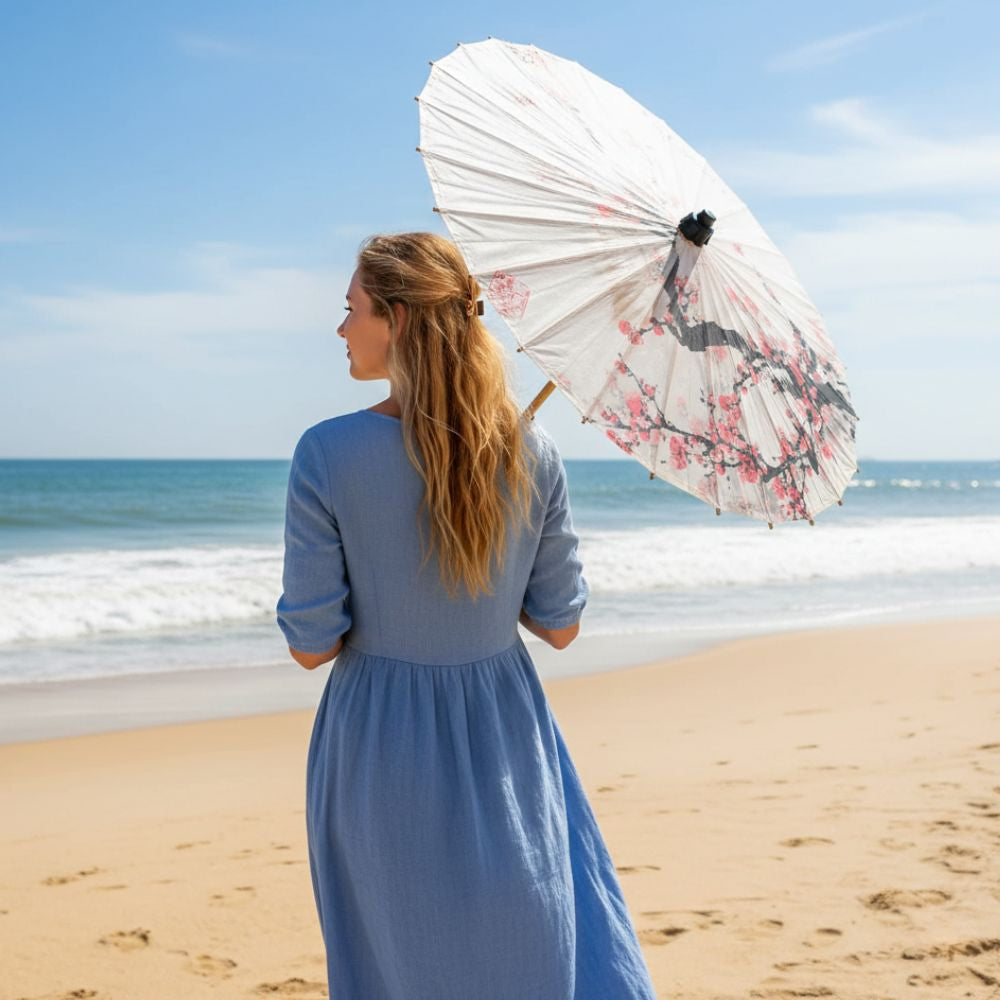 Femme en robe bleue tenant une ombrelle beige ornée de motifs floraux, se tenant sur une plage ensoleillée