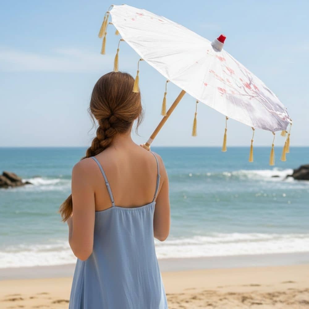 Une femme aux cheveux bruns, portant une robe bleue, tient une ombrelle blanche sur une plage ensoleillée