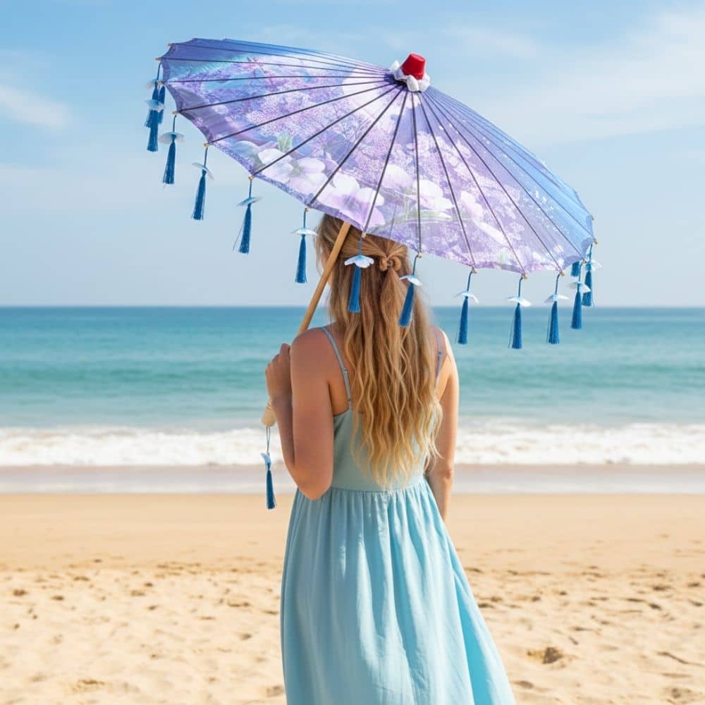 Femme portant une ombrelle pliable bleue avec des franges, debout sur la plage face à la mer