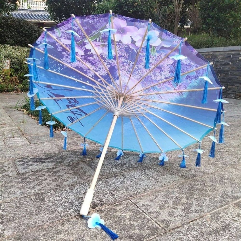 Ombrelle pliable bleu avec motifs floraux, décorée de pompons en tissu, posée sur un sol en pierre