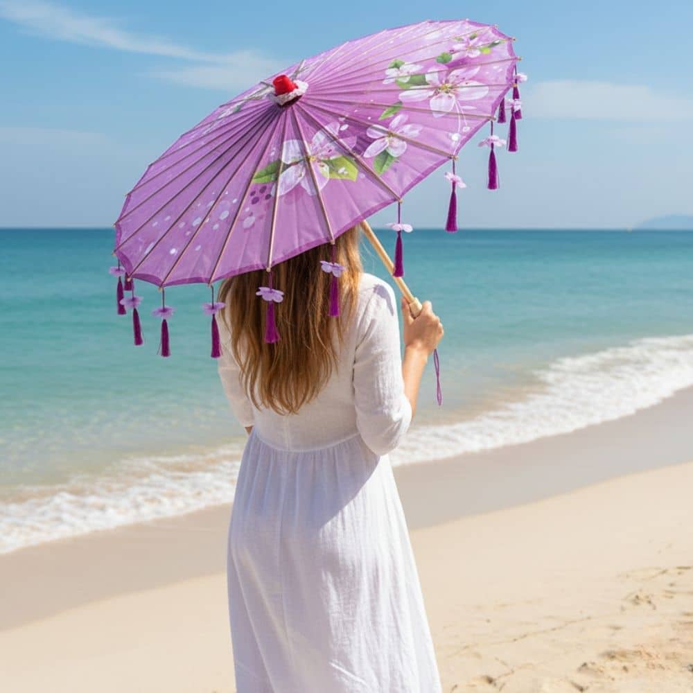 Femme de dos tenant une ombrelle violette décorée, sur une plage avec mer turquoise et ciel dégagé