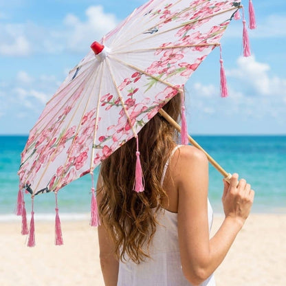 Ombrelle anti-UV rose avec motifs floraux et pompons, tenue légère sur une plage ensoleillée