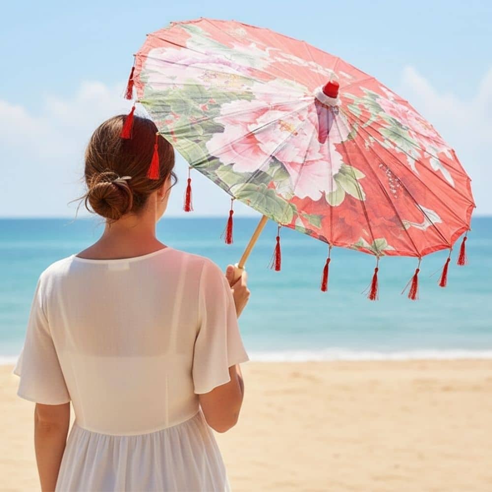 Femme en robe blanche tenant une ombrelle rouge à motifs floraux, face à la mer sous un ciel bleu